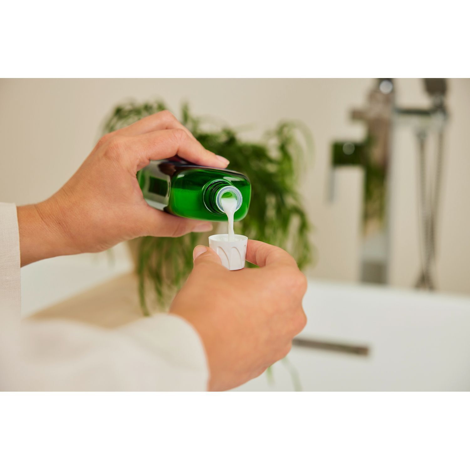 Hände gießen Flüssigkeit aus grüner Flasche in weißen Messbecher. Badezimmerhintergrund mit Waschbecken und Pflanzen.