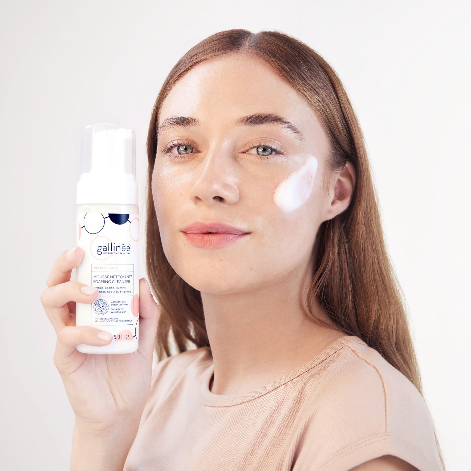 Eine Frau hält eine weiße Flasche mit Pumpverschluss. Aufschrift: gallinée, MOUSSE NETTOYANTE FOAMING CLEANSER. Schaum auf der Wange.