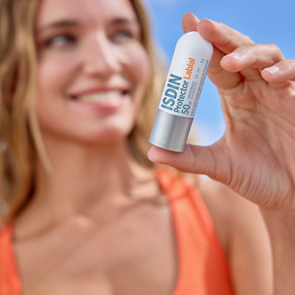 Eine Frau hält einen Lippenbalsam-Stift. Weiß-silberne Hülle mit orangefarbener Schrift: ISDIN Protector Labial 50 SPF. 4g.