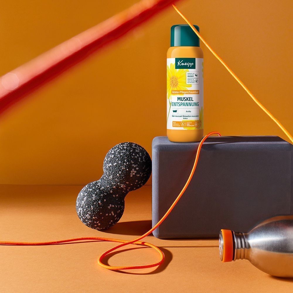 Kneipp® Aroma-Pflegeschaumbad Muskel Entspannung. Gelbe Flasche mit grünem Deckel. Auf orangefarbenem Hintergrund mit Sportutensilien.