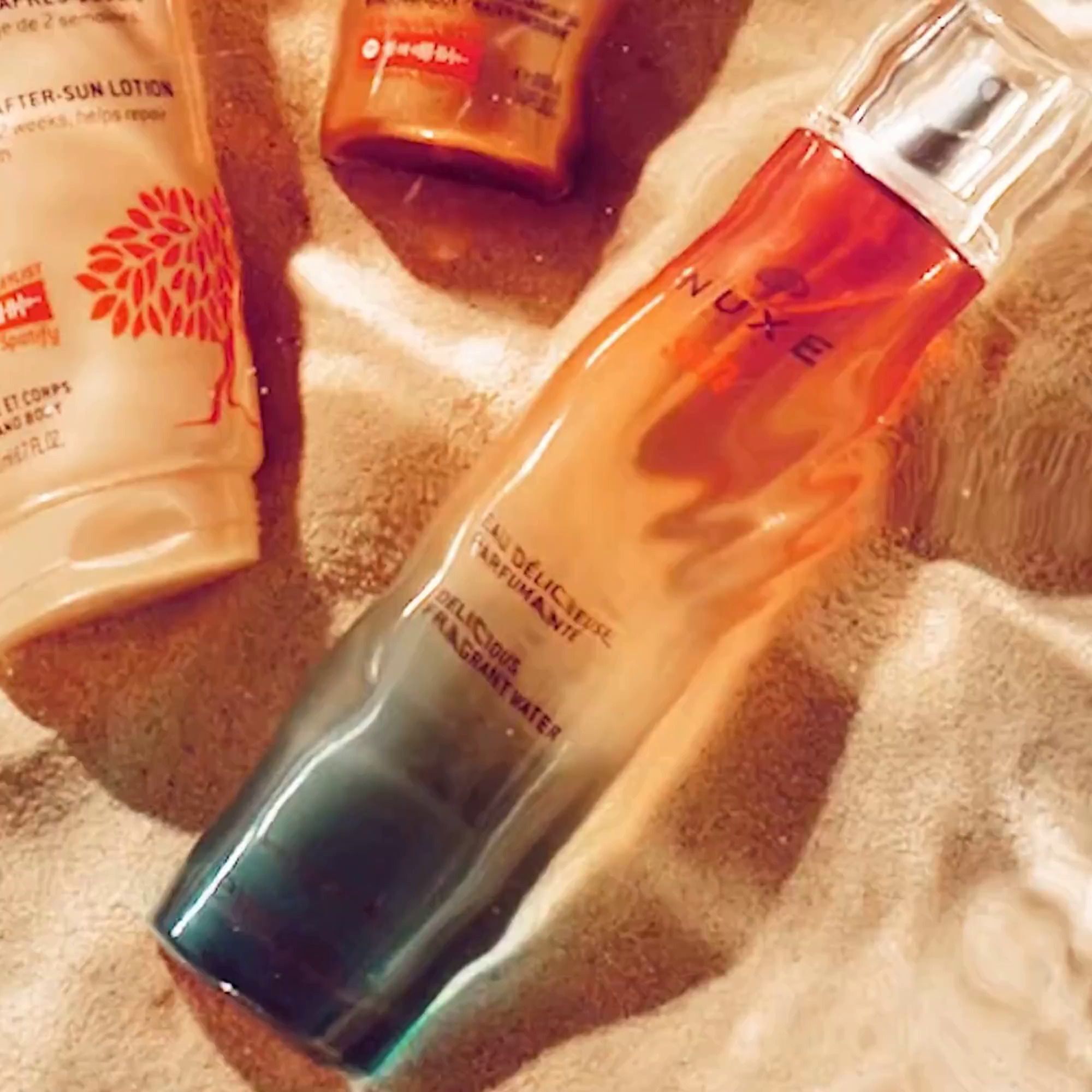 Flakon mit Sprühkopf. Farbverlauf von Blau nach Gelb und Orange. Aufschrift: NUXE Sun, Eau Délicieuse Parfumante, Delicious Fragrant Water. Auf Sand.