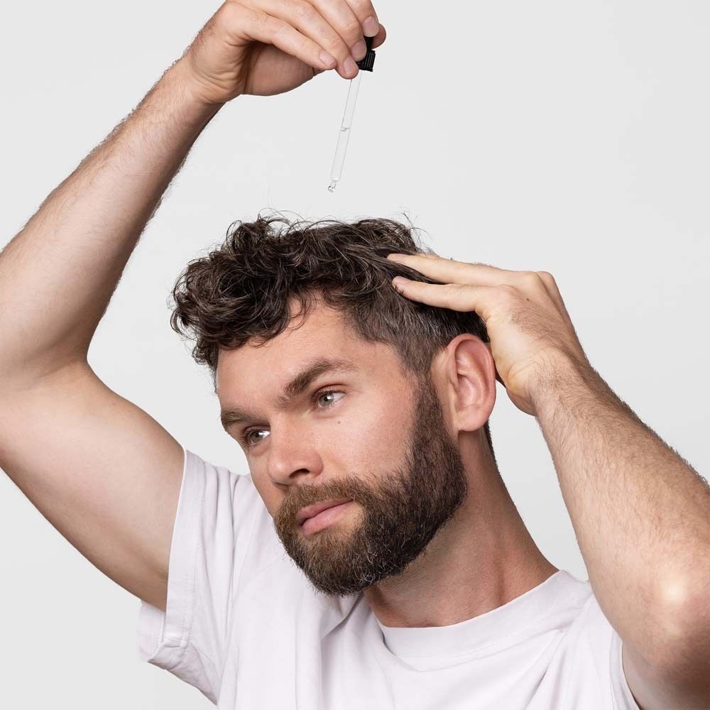 Mann mit Bart hält Pipette über seinen Kopf. Er berührt seine Haare. Weißer Hintergrund.