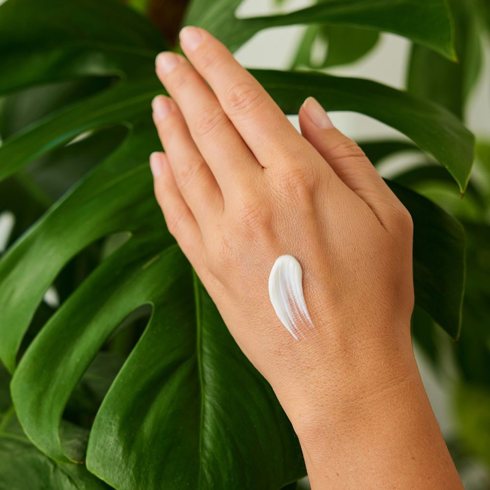 Hand mit einem Klecks Creme. Die Creme ist weiß. Grüner Hintergrund mit Blättern.