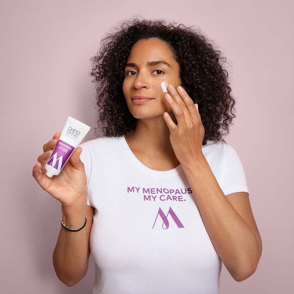 Frau mit lockigem Haar hält eine Tube Creme. Sie trägt ein weißes T-Shirt mit Aufdruck. Hintergrund: Rosa.