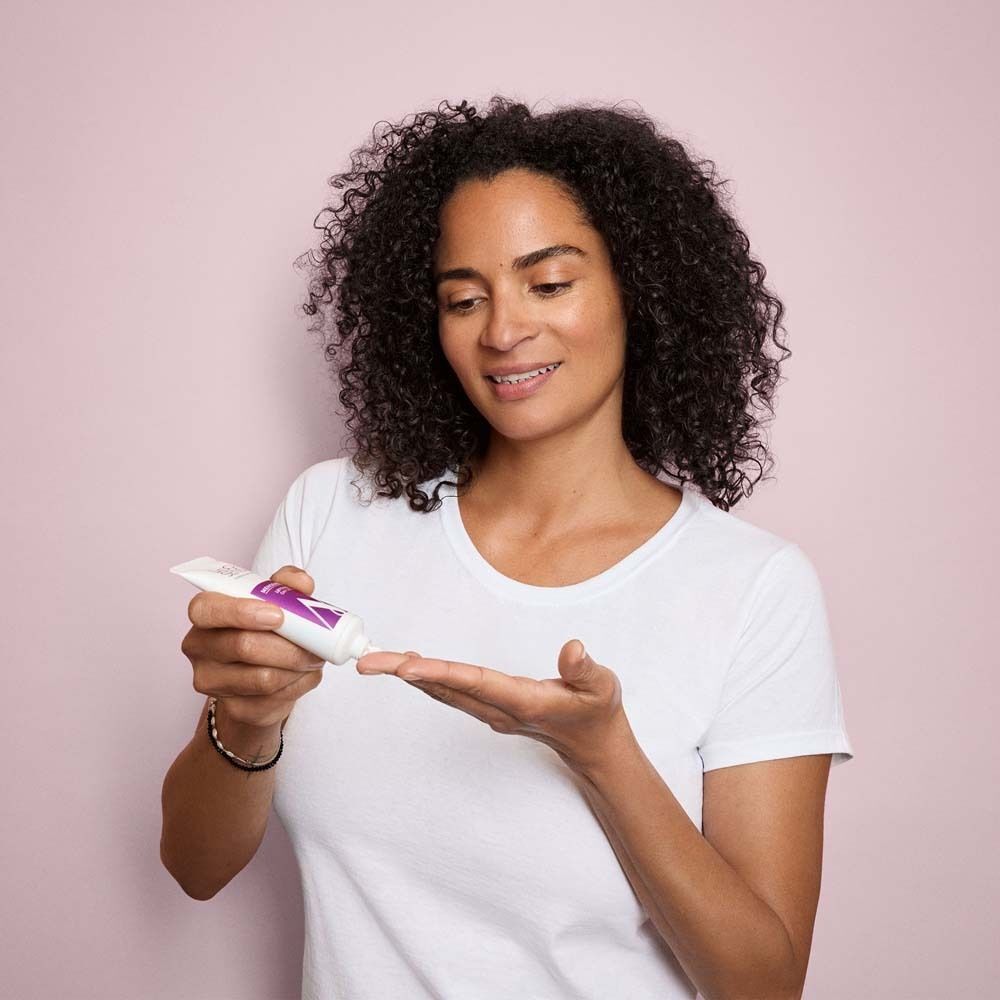 Frau mit lockigem Haar hält eine Tube Creme in der Hand. Sie drückt Creme auf ihre Handfläche. Hintergrund: Rosa.