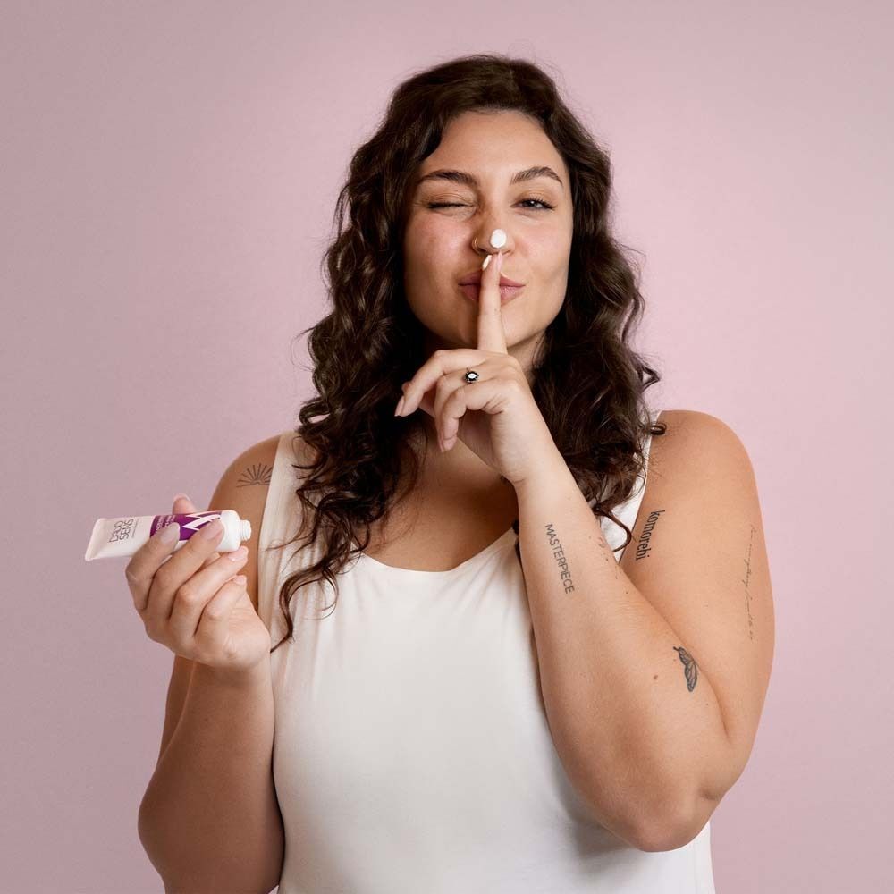 Frau mit lockigem Haar hält eine Tube Creme und legt den Finger auf die Lippen. Hintergrund: Rosa.