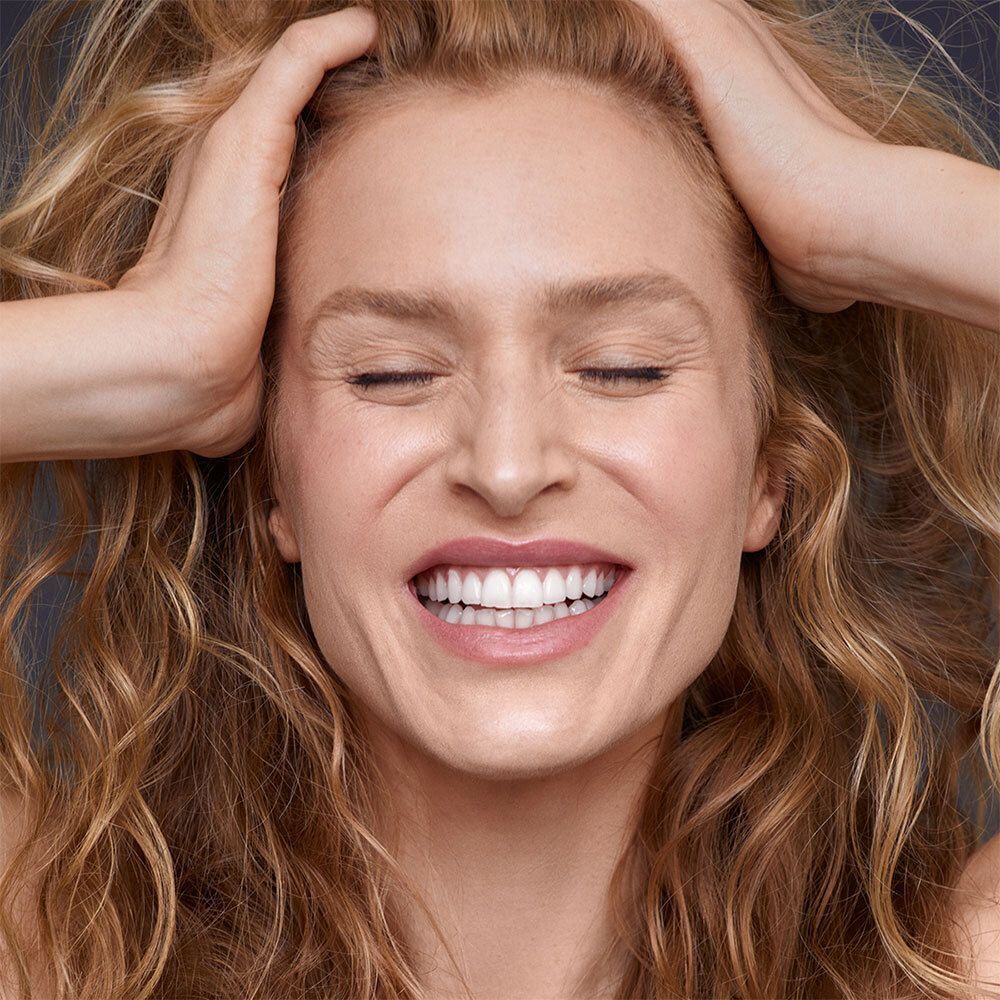 Frau mit blonden Locken, Hände im Haar, Augen geschlossen, lächelnd. Dunkler Hintergrund.