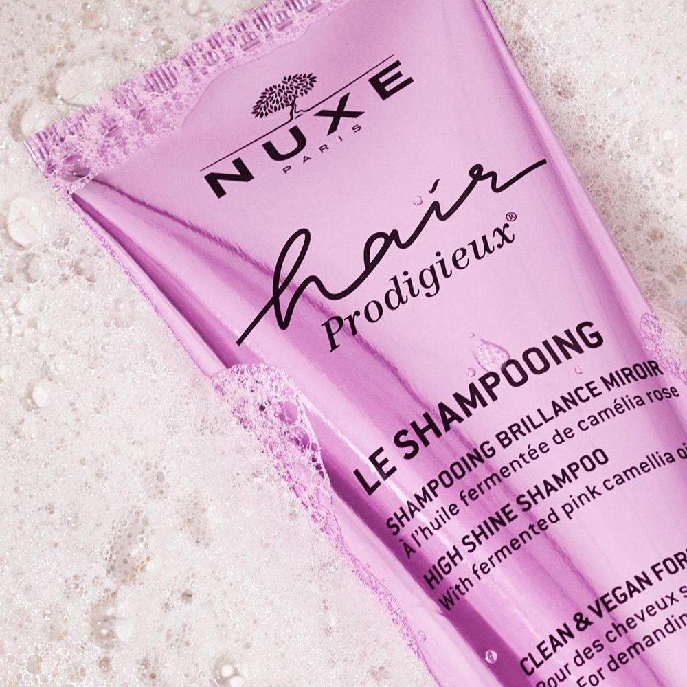 Nahaufnahme einer rosa Shampoo-Tube, umgeben von Seifenschaum. Aufschrift: NUXE, hair Prodigieux, Le Shampooing.