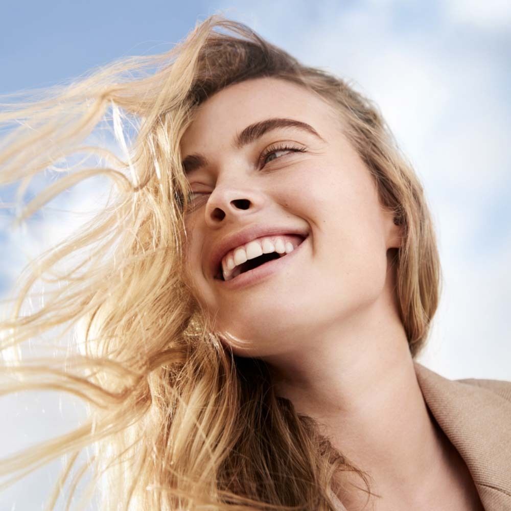 Porträt einer lächelnden Frau mit blonden Haaren. Der Hintergrund ist blau und sonnig.