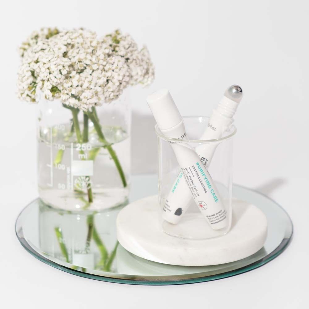 Zwei weiße Roll-on Flaschen in einem Glasbehälter, daneben ein Glas mit weißen Blumen.