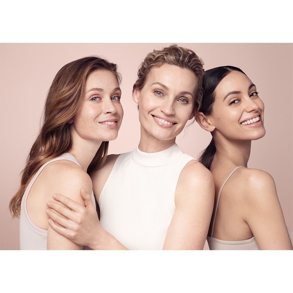 Drei lächelnde Frauen vor rosa Hintergrund. Sie tragen helle Kleidung. Keine Produkte sichtbar.