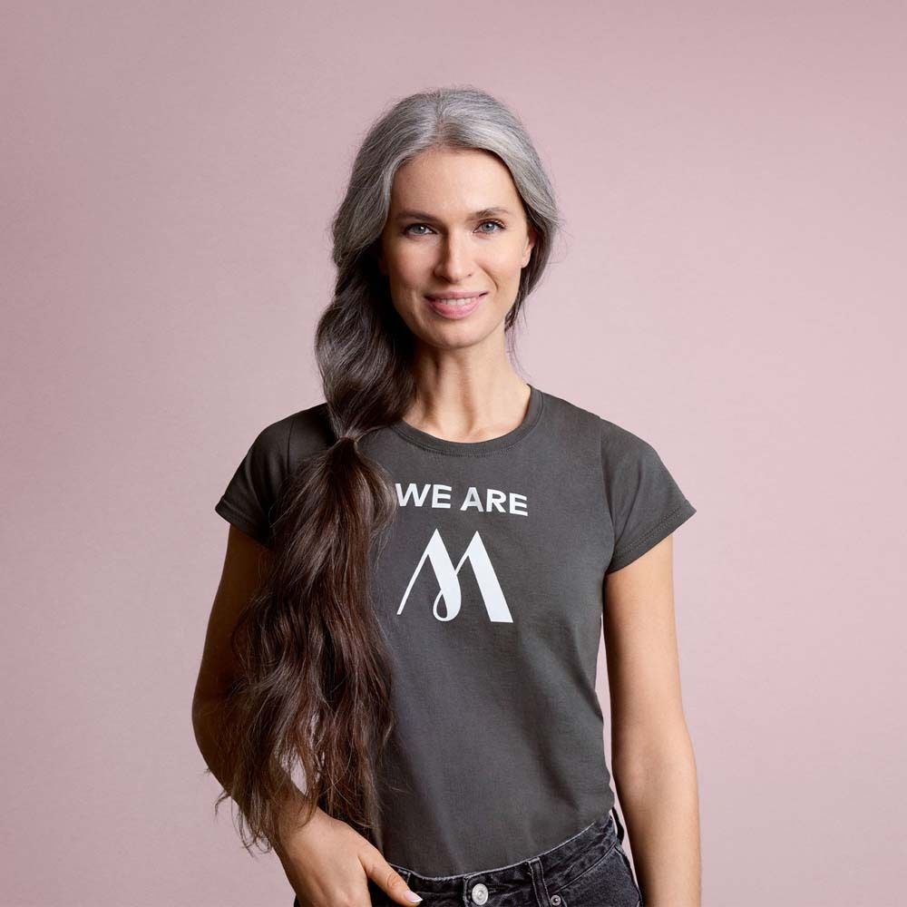 Frau mit grauem Haar trägt graues T-Shirt mit weißem Logo. Text: WE ARE M. Hintergrund: Rosa.