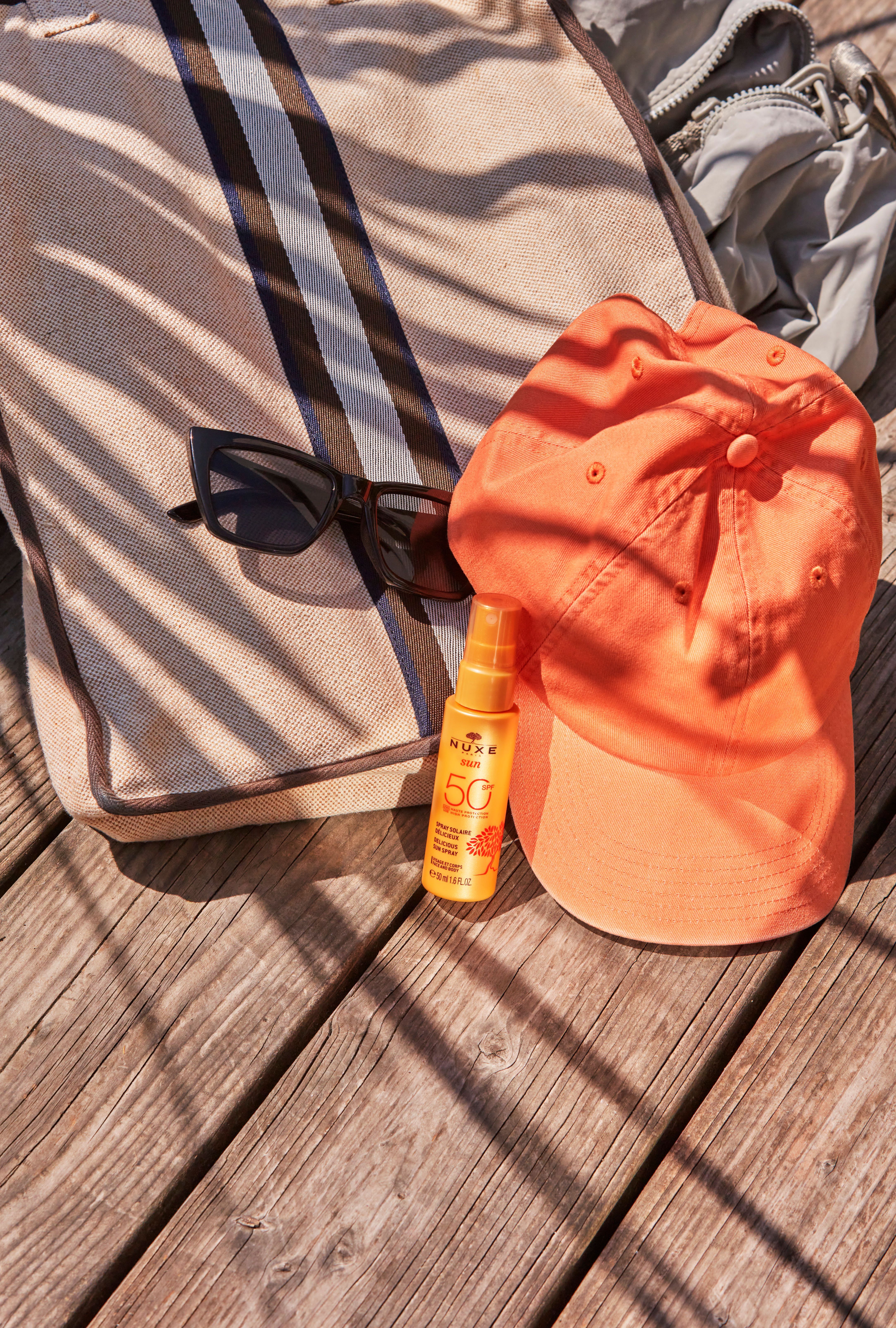 Ein orangefarbenes Sonnenspray neben einer Sonnenbrille, einer orangefarbenen Kappe und einer Strandtasche auf Holz. Aufschrift: NUXE Sun, LSF 50.