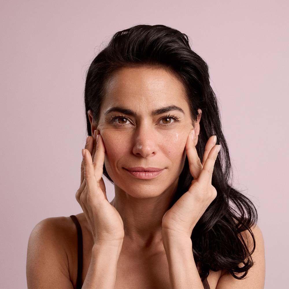 Frau berührt ihr Gesicht. Hintergrund: Rosa. Keine Produkte sichtbar.