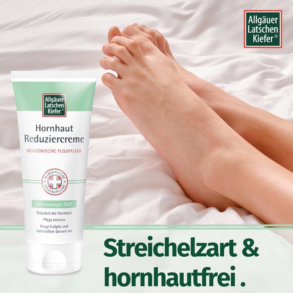 Tube Hornhaut Reduziercreme neben Füßen auf Bettlaken. Weiß-grüne Verpackung mit Logo und Produktinformationen. Text: Streichelzart & hornhautfrei.