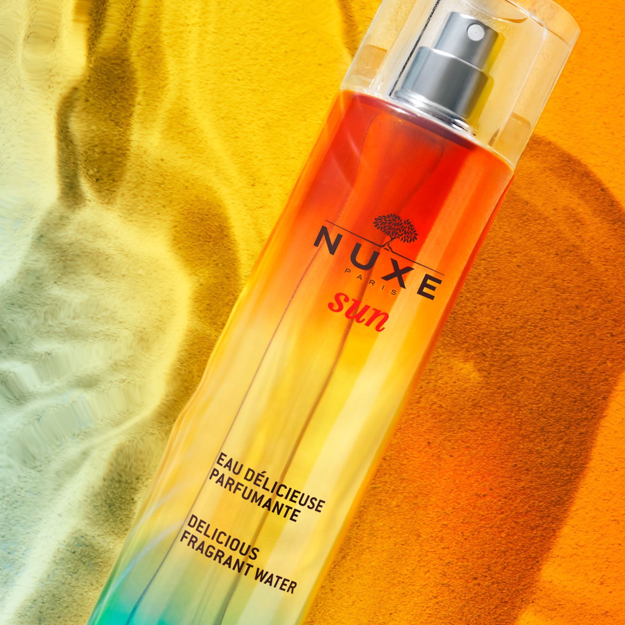 Flakon mit Sprühkopf. Farbverlauf von blau nach orange. Text: NUXE Sun, Eau Délicieuse Parfumante, Delicious Fragrant Water.