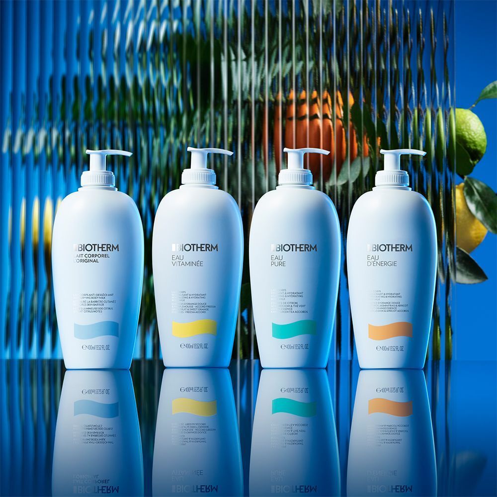 Vier weiße Flaschen BIOTHERM Eau d'Energie Body Milk, jede mit farbigem Akzent. Auf blauem Hintergrund mit Reflexionen.