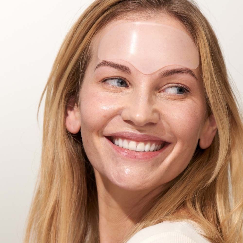 Eine Frau mit blonden Haaren trägt ein transparentes Stirn Pad auf der Stirn. Sie lächelt.