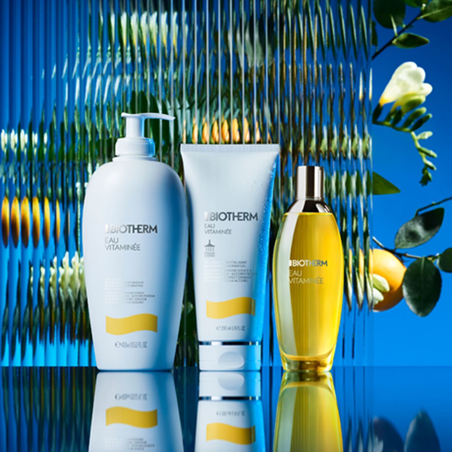 Drei BIOTHERM Eau Vitaminee Produkte auf Glasoberfläche. Links Bodylotion, Mitte Duschgel, rechts Bodyspray.