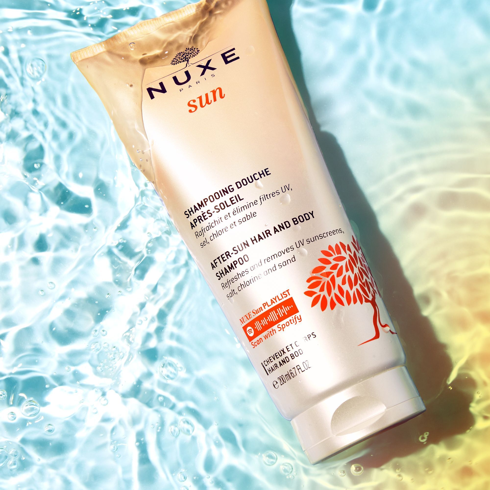 NUXE Sun After Sun Duschshampoo-Tube im Wasser. Weiß-beige Tube mit orangefarbenem Baum-Design. Text: After-Sun Hair and Body Shampoo.