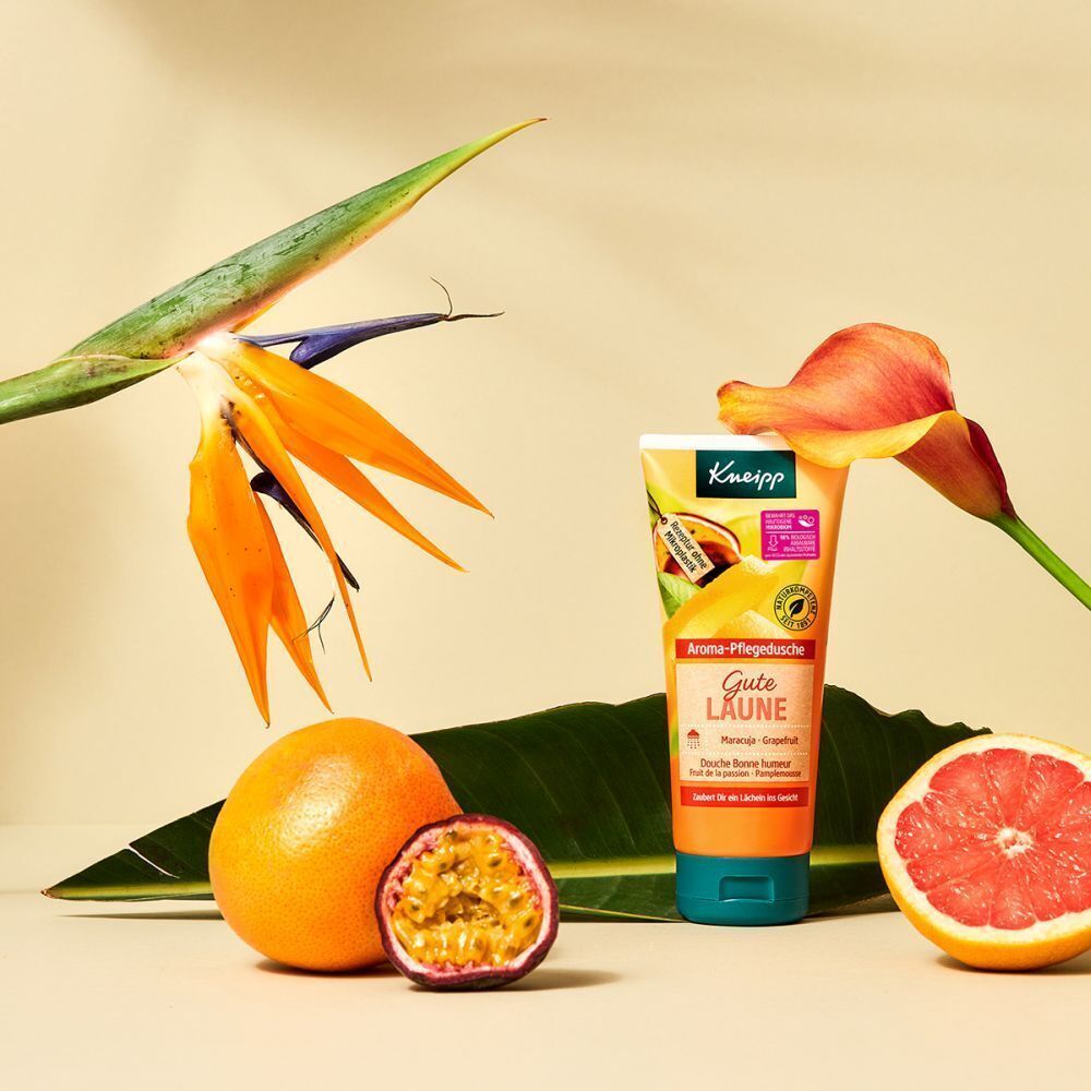 Eine Tube Kneipp® Aroma-Pflegedusche Gute Laune neben Früchten und Blumen. Maracuja, Grapefruit, orangefarbene Blüten und grünes Blatt.