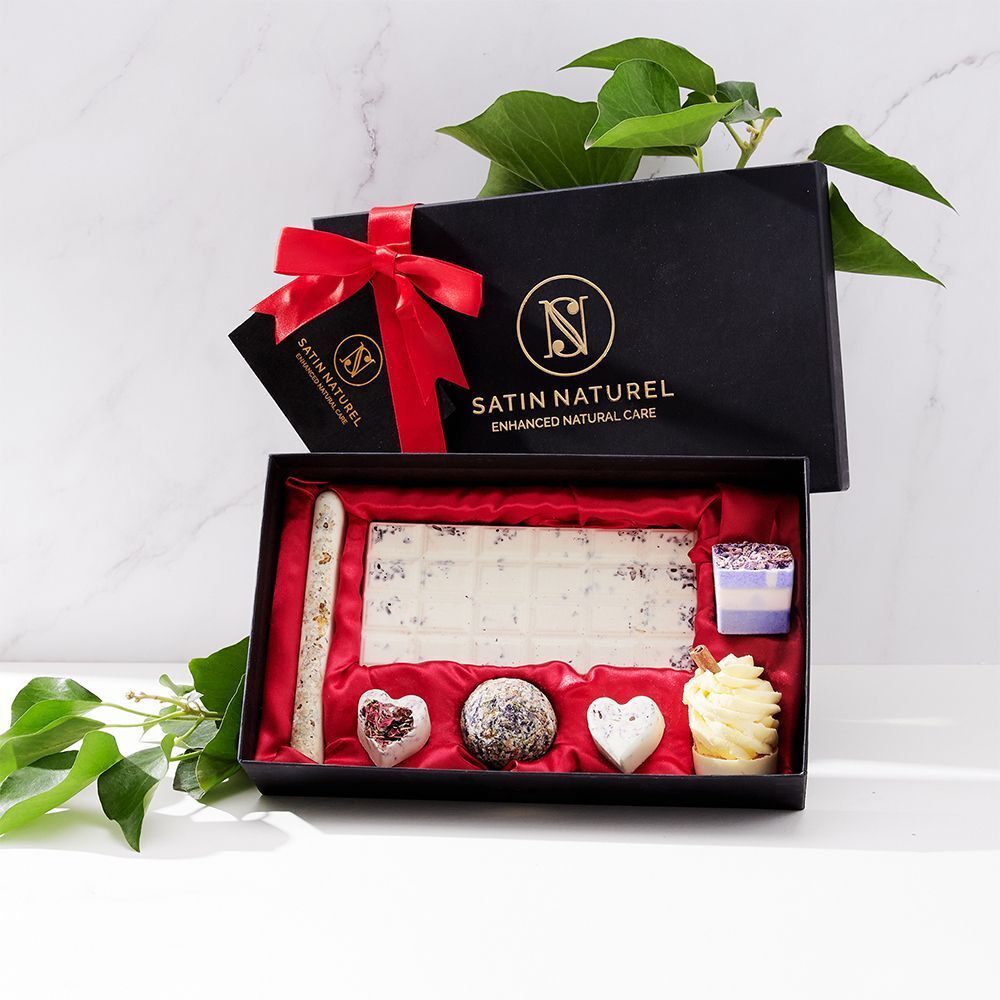 Geöffnete schwarze Geschenkbox mit roten Innenfutter. Enthält Badepralinen. Logo: SATIN NATUREL. Grüne Blätter im Hintergrund.