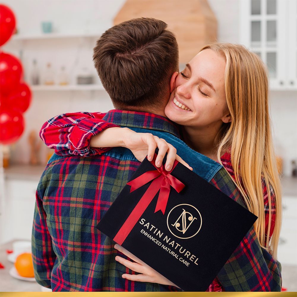 Personen umarmen sich. Eine Person hält eine schwarze Geschenkbox mit roter Schleife. Logo: SATIN NATUREL. Text auf der Box.