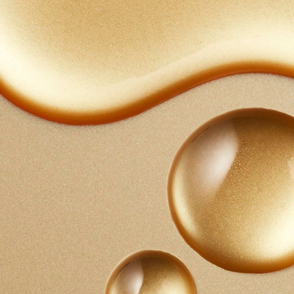 Nahaufnahme von goldenen Flüssigkeitstropfen auf einer strukturierten Oberfläche. Runde und ovale Formen.