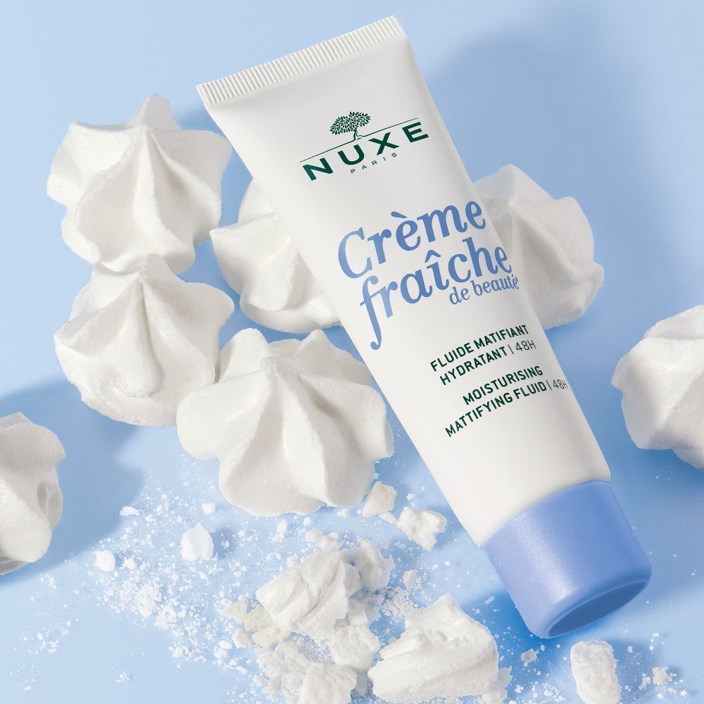 Weiße Tube mit blauer Kappe, umgeben von weißen, meringue-artigen Formen. Aufschrift: NUXE, Crème fraîche de beauté.