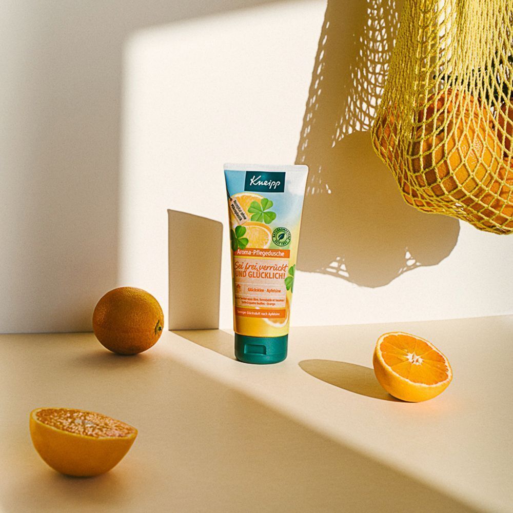 Tube Kneipp Aroma-Pflegedusche neben Orangen und einer Netztasche. Produkt mit Kleeblatt- und Orangendesign. Aufschrift: Sei frei, verrückt und glücklich!