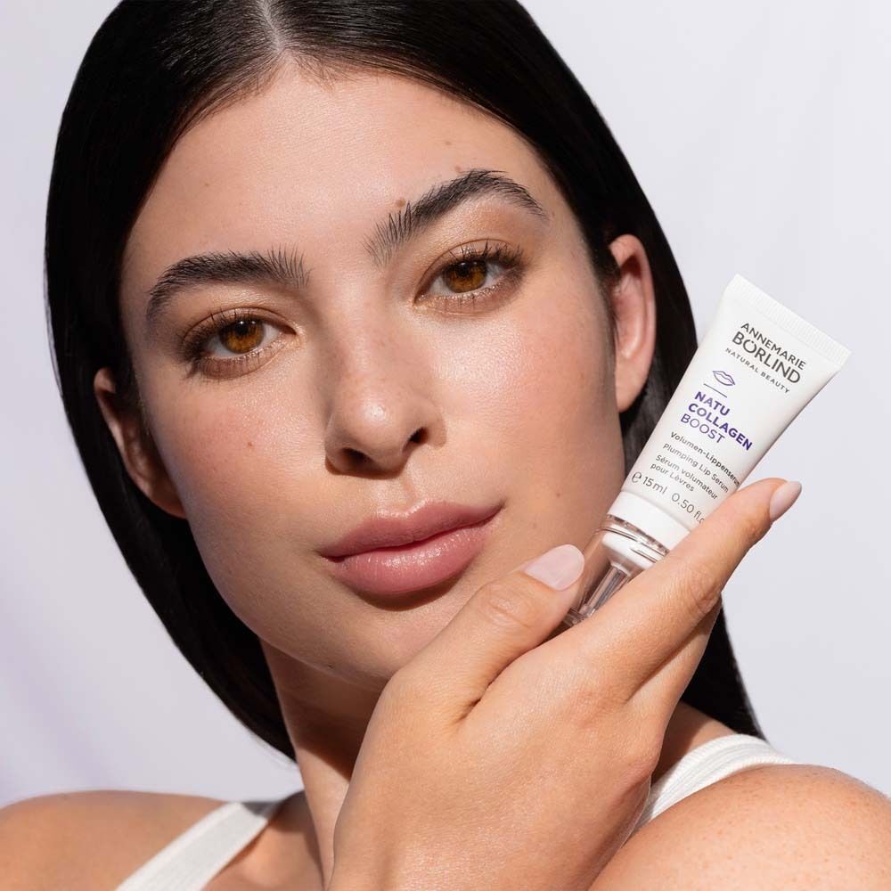 Eine Frau hält eine Tube ANNEMARIE BÖRLIND Natu Collagen Boost Volumen-Lippenserum neben ihr Gesicht.
