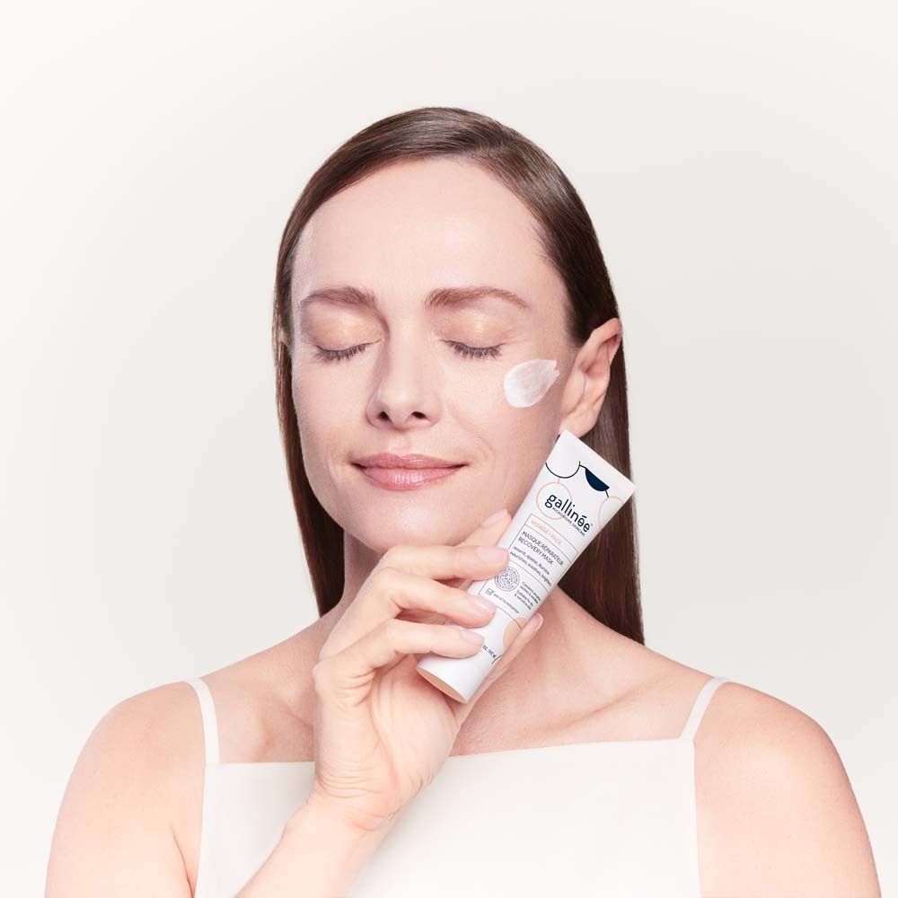 Frau hält eine Tube Gesichtsmaske. Sie hat die Augen geschlossen und trägt ein weißes Top. Auf der Haut ist Creme.