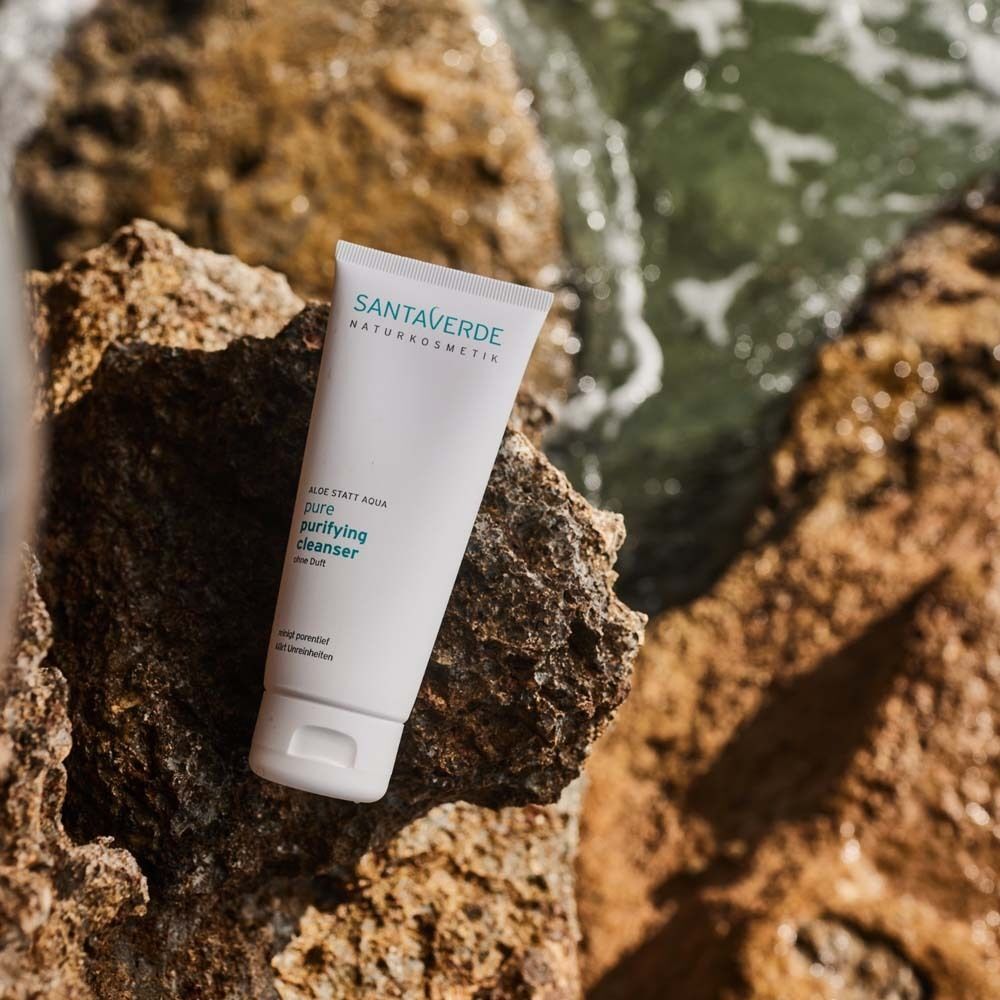 Eine Tube SANTAVERDE pure purifying cleanser auf einem Felsen. Hintergrund: Meerwasser. Produktname und weitere Informationen sind aufgedruckt.