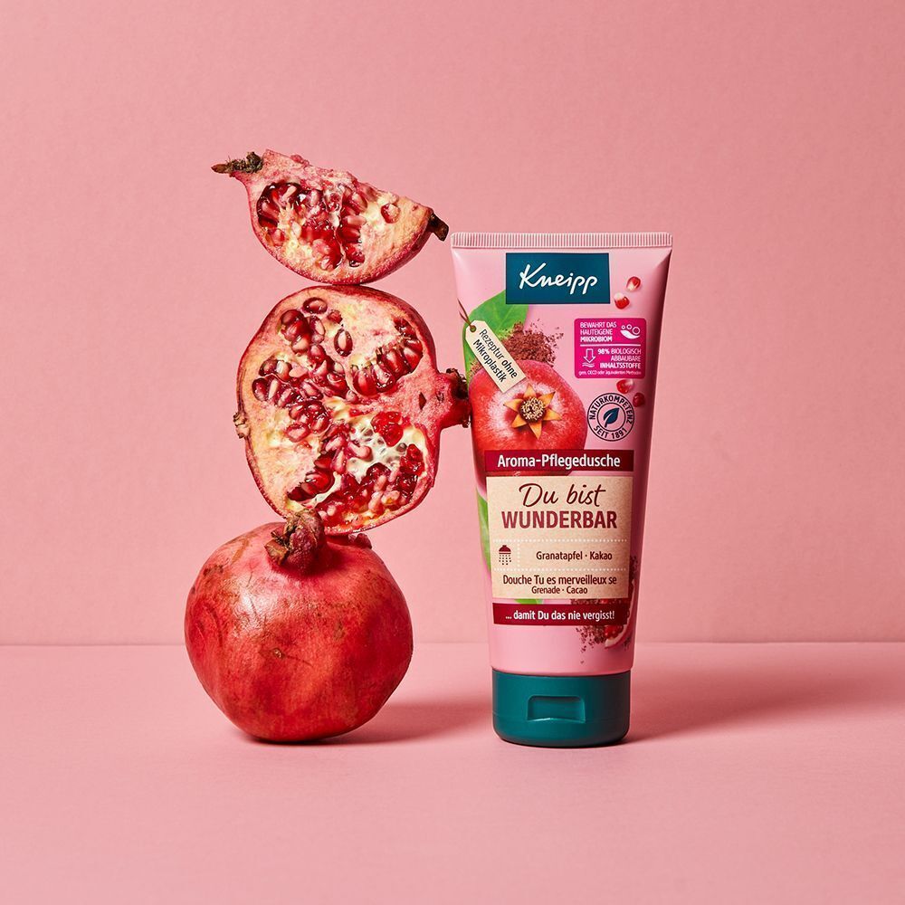 Eine Kneipp® Aroma-Pflegedusche-Tube neben Granatäpfeln. Rosa Tube mit Text: Du bist wunderbar. Hintergrund rosa.