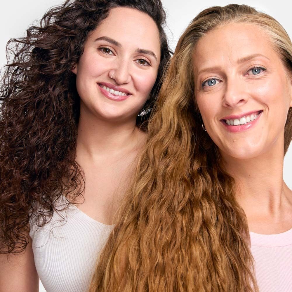Zwei Frauen mit lockigem und gewelltem Haar. Eine Frau mit braunen Locken, die andere mit blondem, gewelltem Haar. Beide lächeln.