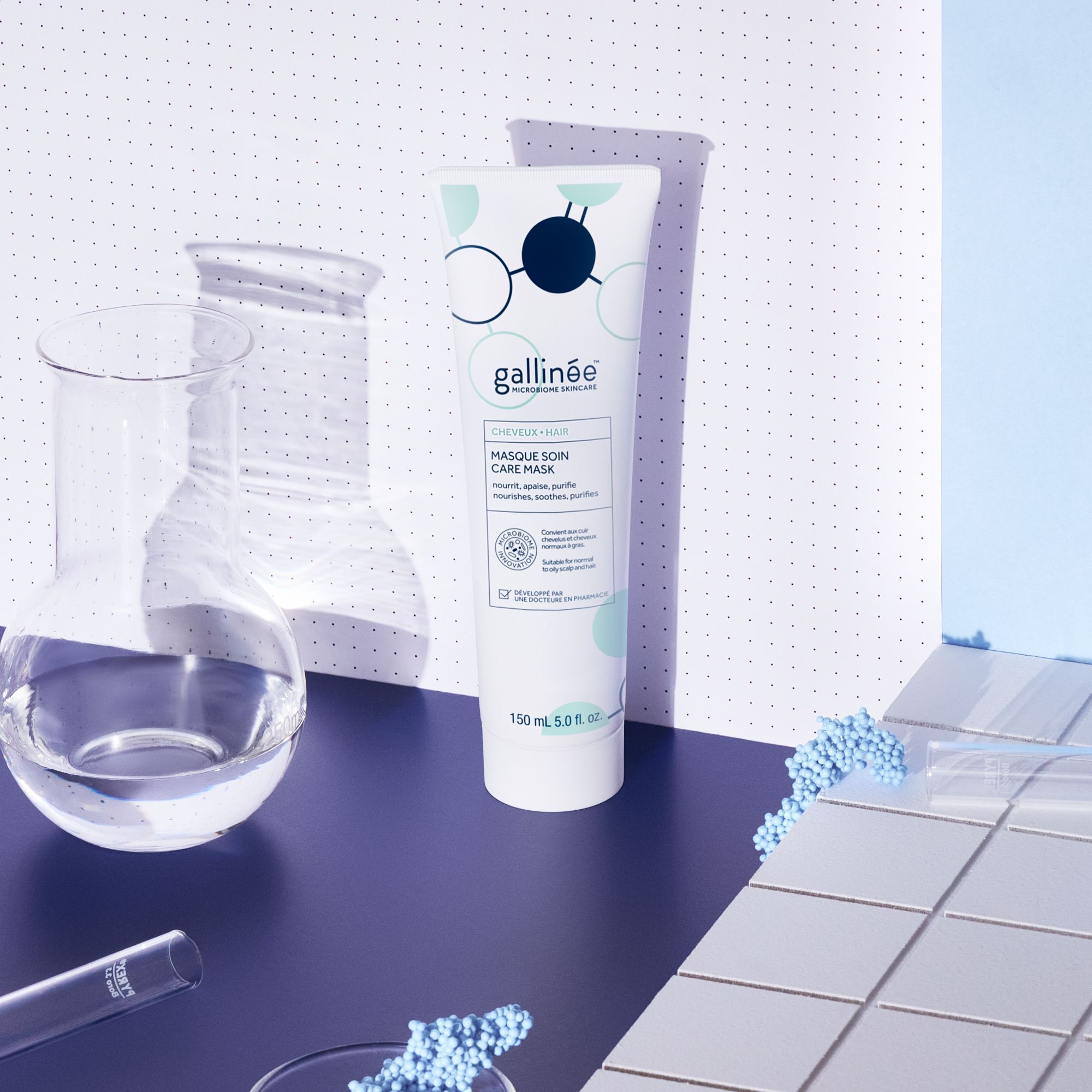 Eine Tube Haarmaske steht vor einem Laborhintergrund. Marke: gallinée. Aufschrift: Masque Soin Care Mask. 150 ml. Blaue Kügelchen und Glaswaren.