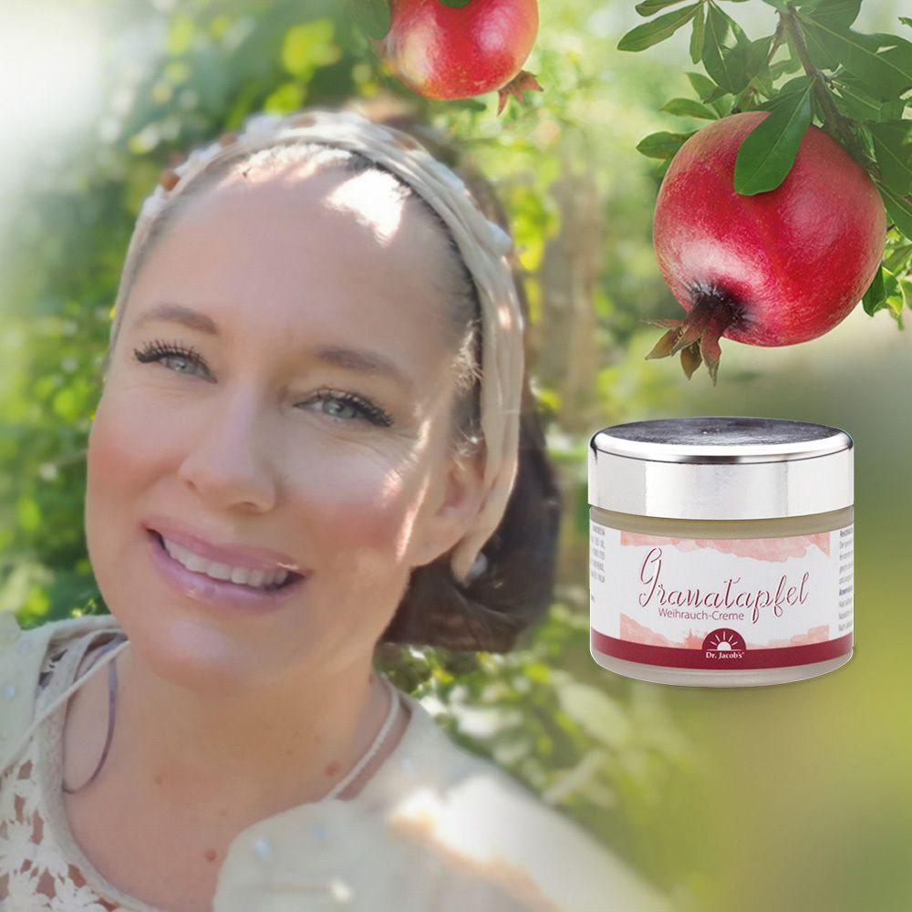 Frau mit Stirnband lächelt. Neben ihr ein Tiegel Granatapfel-Weihrauch-Creme. Hintergrund: Granatapfelbaum mit Früchten.