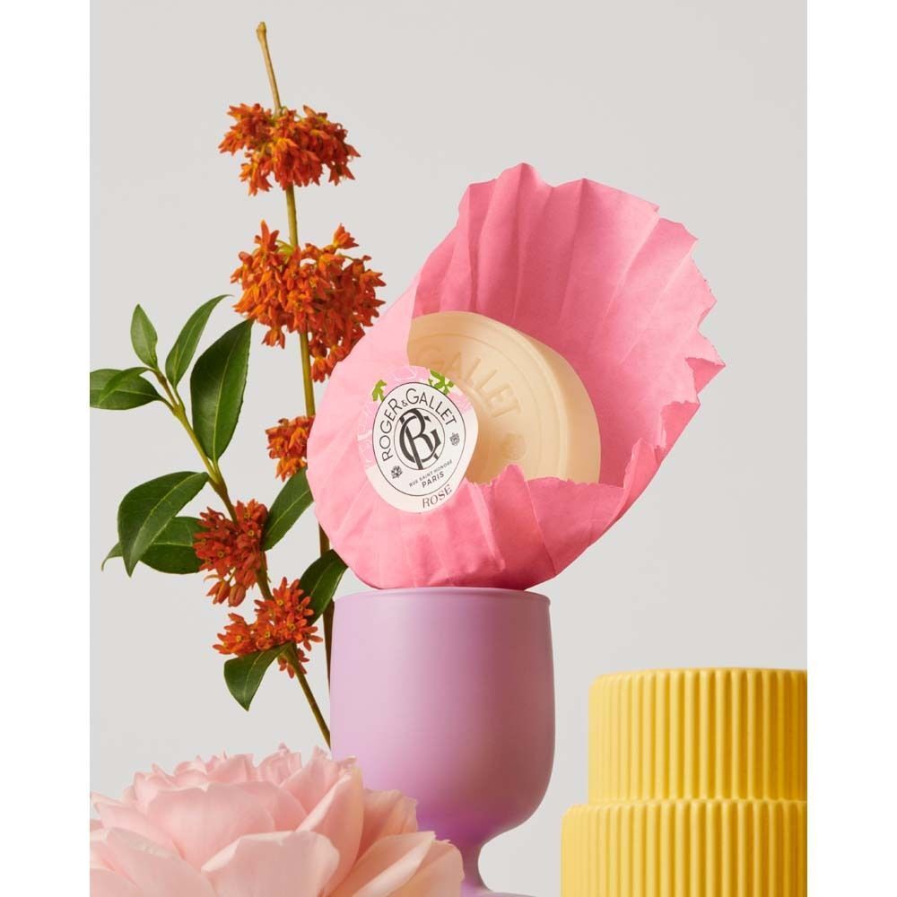 Runde, cremefarbene Seife in rosa Papierverpackung, platziert in einem lila Becher. Dekoration mit Blumen.