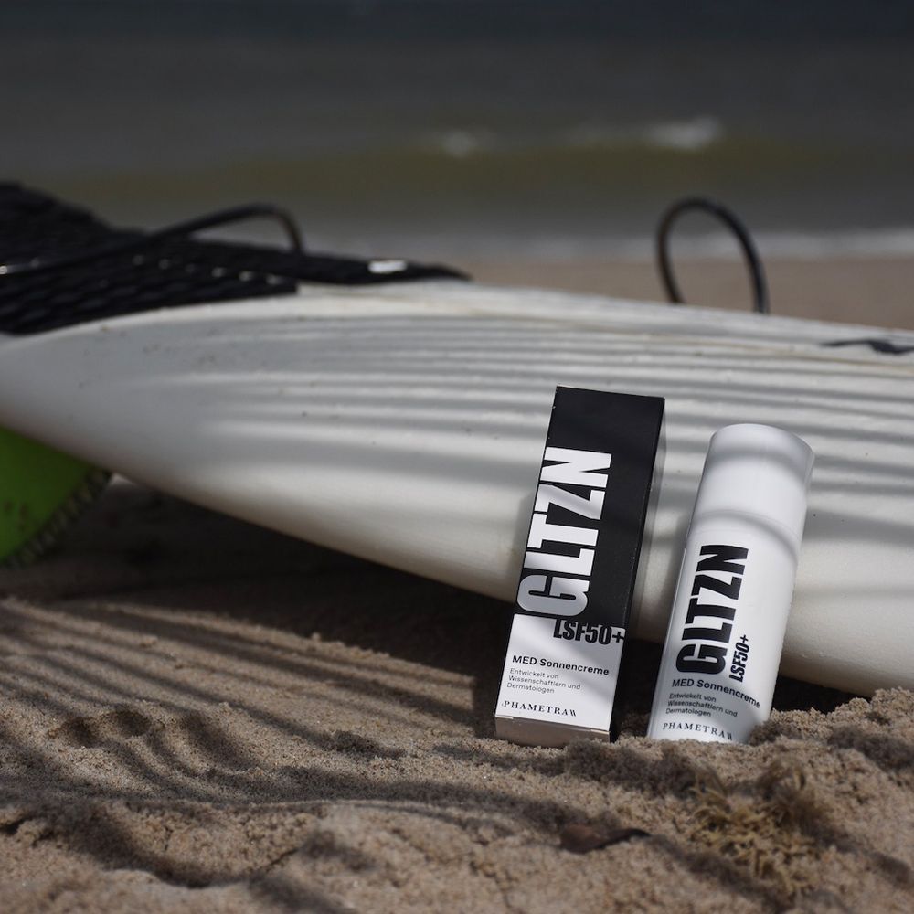 GLTZN MED Sonnencreme LSF 50+ Flasche und Verpackung am Strand neben einem Surfbrett.