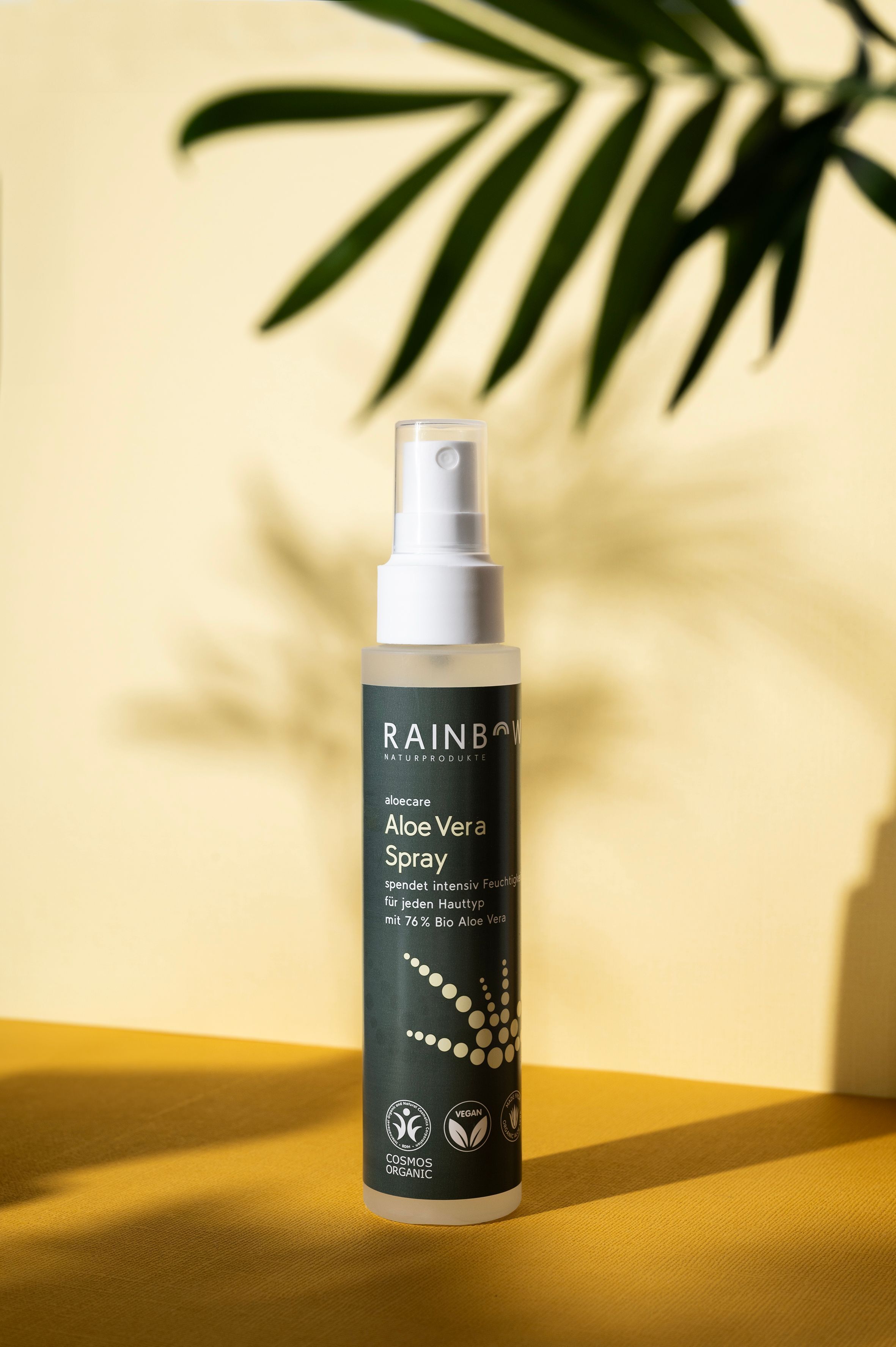 Aloe Vera Spray auf gelbem Untergrund. Grüne Flasche mit weißem Sprühkopf. Vegan- und Cosmos Organic-Siegel.