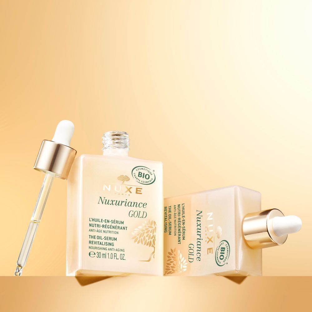 Zwei quadratische Flakons NUXE Nuxuriance Gold Öl-Serum. Goldfarbene Deckel mit Pipetten. Aufschrift: Nuxuriance GOLD, L'HUILE-EN-SÉRUM, e 30 ml. Hintergrund: goldfarben.