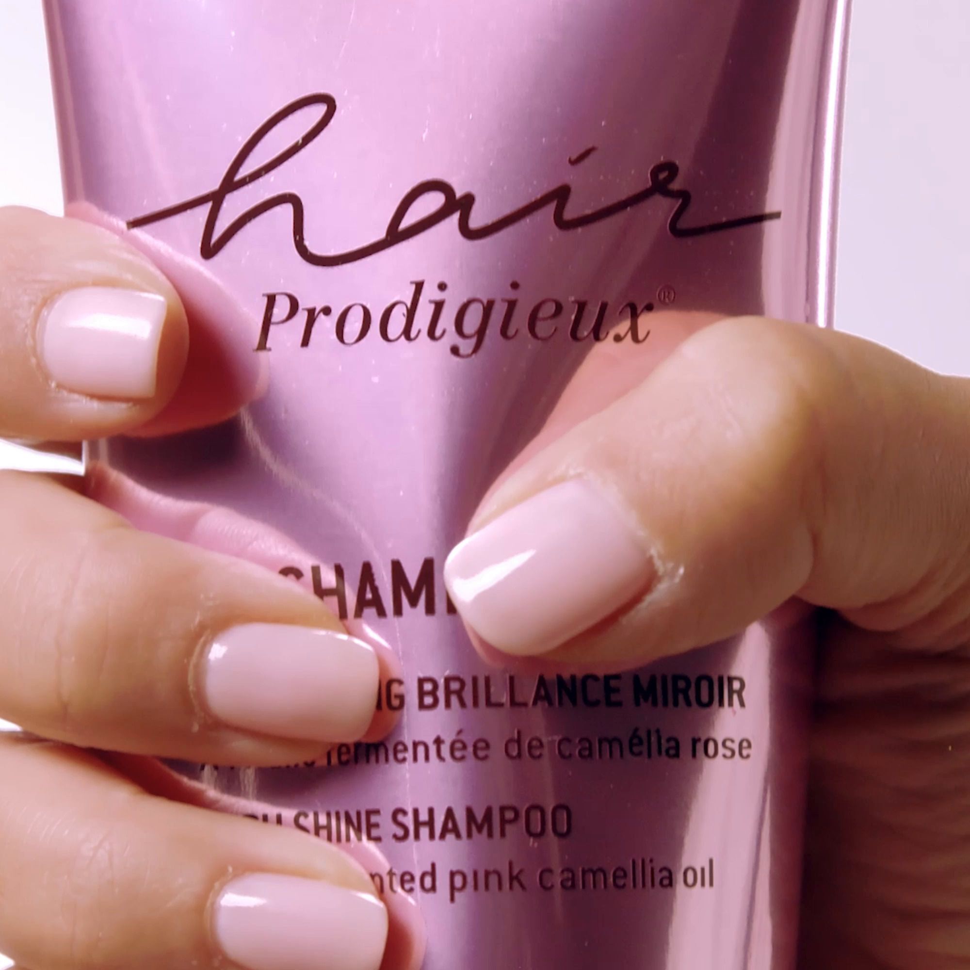 Nahaufnahme einer Hand, die eine rosa Shampoo-Flasche hält. Aufschrift: Hair Prodigieux, Shampooing Brillance Miroir.