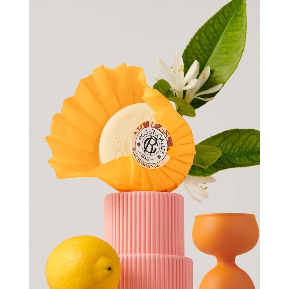 Runde, weiße Seife in gelber Papierverpackung, dekoriert mit Zitrone, Blüten und einem rosa Podest.