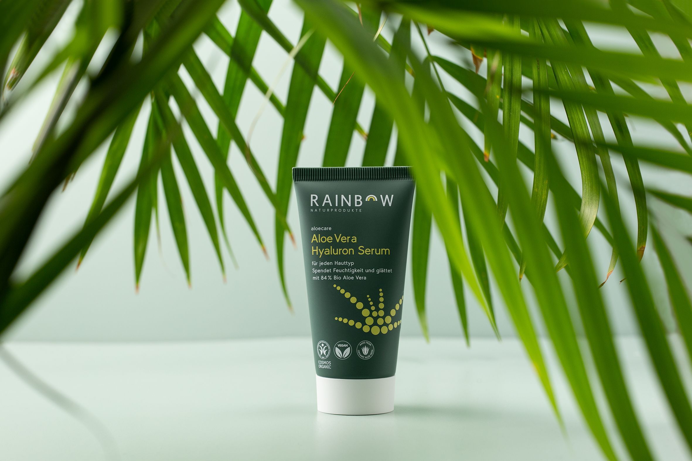 Tube Aloe Vera Hyaluron Serum. Grün-weiße Verpackung. Produktname und Logos. Vegan- und Cosmos Organic-Siegel. Vor Palmenblättern.