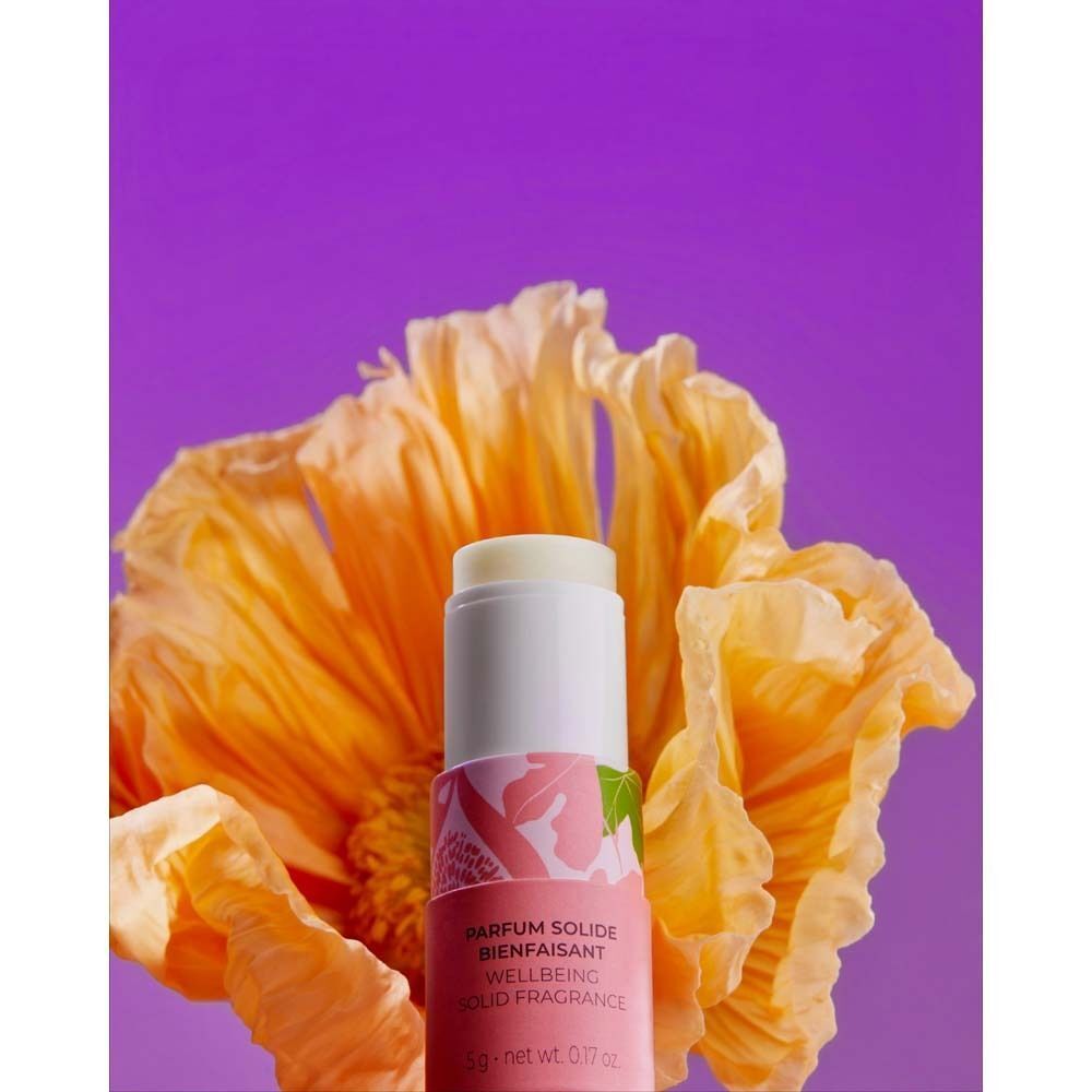 Parfumstick vor gelber Blüte. Weißer Deckel, rosafarbener Boden. Aufschrift: PARFUM SOLIDE BIENFAISANT WELLBEING SOLID FRAGRANCE.