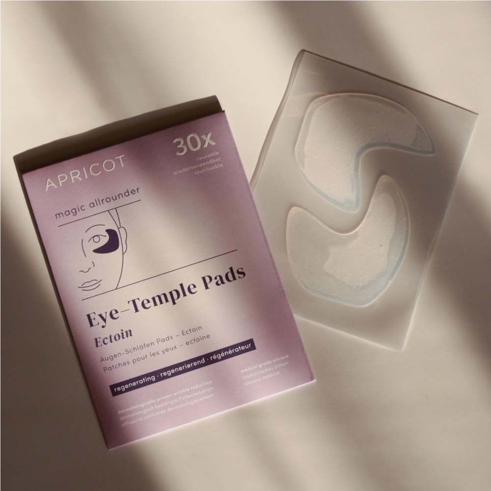 Packung Augen-Schläfen Pads und daneben liegende Pads. Auf der Packung steht "APRICOT", "30x" und "Eye-Temple Pads Ectoin".