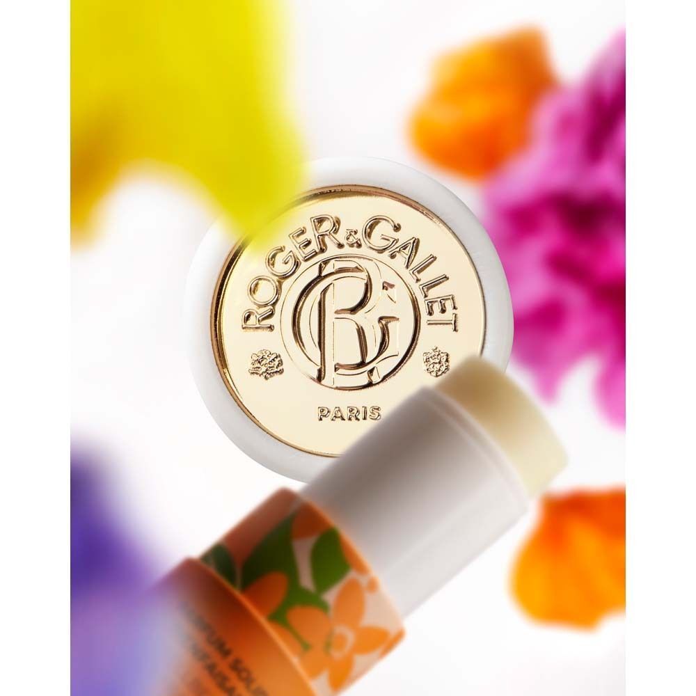 Parfumstick mit goldenem Deckel und floralem Muster. Weißer Deckel, Text: ROGER & GALLET PARIS. Blüten im Hintergrund.