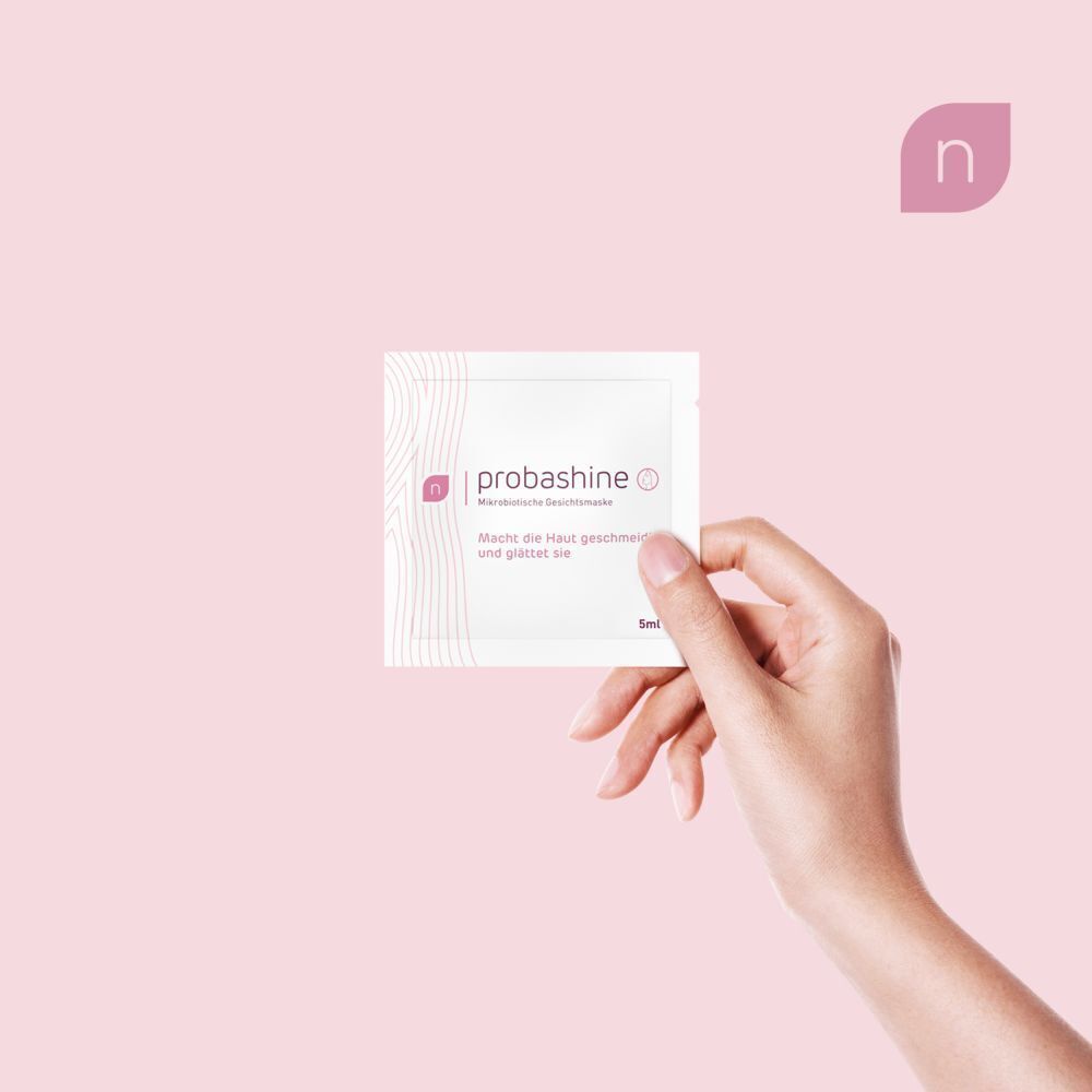 Hand hält einen nupure probashine Maskenbeutel. Rosa Hintergrund. Text: Macht die Haut geschmeidig.