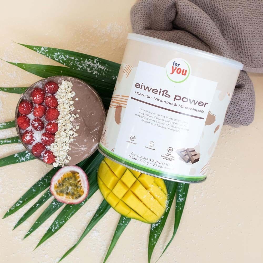 Dose "for you eiweiß power Chocolat noir" neben Smoothie mit Himbeeren und Mango. Dekoration mit Palmblatt und Handtuch.