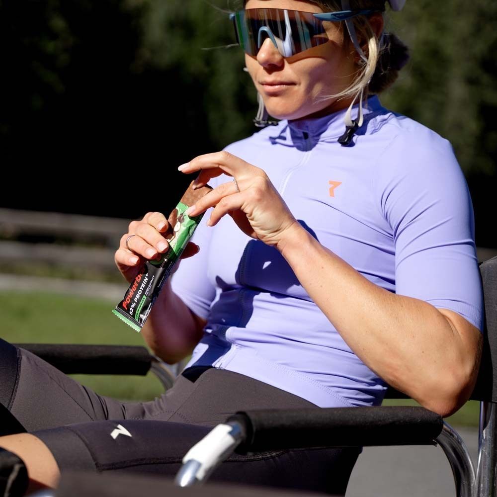 Eine Frau in Sportkleidung hält eine PowerBar 52% Protein Riegel. Sie sitzt in einem Rollstuhl und trägt eine Sonnenbrille.